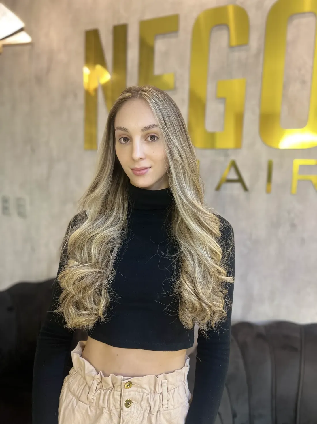 Cliente loira com mechas iluminadas, painel dourado Nego Hair ao fundo